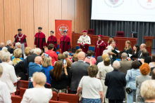 A ceremony in the auditorium. Seven people on stage: rector and dean authorities in togas and birettas, one holding an open book. In the background is a red banner with the faculty emblem. In the foreground, participants take pictures of the authorities.