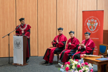 A man in a red and black toga - the dean of WL is speaking at a lectern with the university's logo. Next to him sit three people in togas, clapping. In the background is a red banner with the logo of the Faculty of Medicine. In the foreground is a table with a bouquet of flowers.