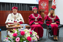 Three people in academic togas sit on chairs. The person on the left in a white toga with a scepter, the others in red togas and birettas. In front of them is a table with a bouquet of flowers. In the background is a banner with the university's coat of arms and the inscription “Veritatis Splendor.”