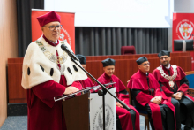The rector, in red ceremonial dress with a white collar and rector's chain, speaks into the microphone. In the background are four people in red togas and birettas, two red banners with the university and department logos, and a wooden lectern.