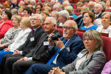 A group of people are sitting in an auditorium during a conference. In the foreground a man in a blue suit is holding a camera, next to him a woman in a plaid jacket. Other participants in red chairs are visible in the background. They are listening intently.