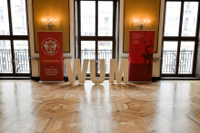 An elegant room with a wooden floor and large windows. In the centre stand the large white letters ‘WUM’. On the sides two red roll-ups with the inscription ‘Warsaw Medical University Faculty of Medicine’. On one the university's coat of arms, on the other a graphic of a chalice.
