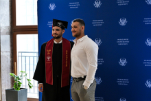 Two men pose against a blue wall with the logo of the Warsaw Medical University. One is wearing a toga, black biretta and maroon sash with the words “Class of 2023” and “Graduate”. The other has a white shirt and grey trousers. In the background is a window and a green plant in a pot.
