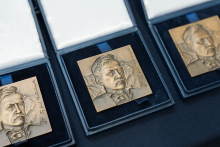 Three gold medals of square shape, housed in open black boxes with white interior. Each medal shows a relief with a face and engraved text on the right side.