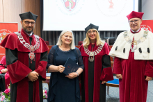 Four people in academic attire, three in red togas with chains, one in a white toga. In the center, a woman in a navy blue dress holds a document. In the background are university logos and curtains.