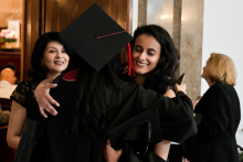 Two people hug during a graduation ceremony. One is wearing a black toga and a square cap with a red tang. The other holds a pink diploma and phone. In the background, other participants, including a man bent over a table. Bright interior with wooden floor and wall sconces.