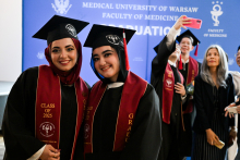 A group of female graduates of the Faculty of Medicine at Warsaw Medical University. In the foreground two people wearing togas, birettas and maroon sashes with the inscription ‘CLASS OF 2025’ and ‘GRADUATE’. In the background, a blue banner with the words “MEDICAL UNIVERSITY OF WARSAW FACULTY OF MEDICINE GRADUATION”. Others take a selfie.