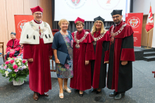 Five people pose for a group photo. Four in red togas with chains, one in a colorful dress holding a blue briefcase. In the background are red banners with the university's coat of arms, a screen with the logo and the words “Universitas Medica.” On the floor a bouquet of flowers in a white basket.