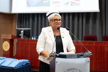 Uroczystość czepkowania absolwentów pielęgniarstwa i położnictwa. Kobieta przemawia przy mównicy. Kobieta ubrana jest w biały żakiet, czarną sukienkę i czepek pielęgniarski.