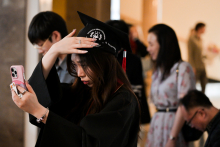 A young woman dressed in a black toga improves her biretta while looking at her mobile phone. In the background you can see other people, participants in the event.