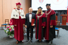 Four people stand against a wooden wall and screen. Three in red and black togas with chains, one in a suit holds a red briefcase. On the left is a bouquet of flowers, on the right a lectern with the inscription “Veritas Splendor.”