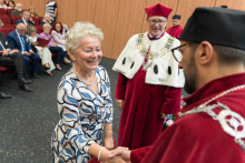  An elderly woman in a dress with blue and white waves and pearls squeezes the hand of a man in red academic attire. In the background, a second man in a red toga with a fur collar and rector's chain. Behind them sit formally dressed guests.