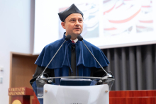 Uroczystość czepkowania absolwentów pielęgniarstwa i położnictwa.  Mężczyzna stoi przy mównicy, przemawia do publiczności. ubrany jest w niebieską togę i biret.