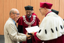An older man in a light-colored suit receives a document from a person in a white coat and red biretta. In the background, a person in a red academic outfit with a chain. Behind them is a wooden panel wall.