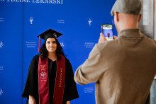 A graduate in a black toga and biretta poses for a photo against a blue wall with the words “Faculty of Medicine” and “Warsaw Medical University” written on it. She has a maroon sash with the inscriptions “Class of 2025” and “Graduate”. In front of her stands a photographer with a phone.
