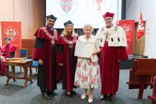 Four people, including three wearing academic togas and birettas. In the center, a woman in a white floral dress holds a diploma. In the background are two banners and a screen with the university's logo. In the foreground on the left a table and a chair.