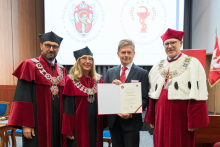 Four people stand side by side, three wearing maroon academic togas with chains. The man in the middle holds a diploma, he is dressed in a suit and red tie. In the background is a screen with the medical school's logo and a flag with a coat of arms.