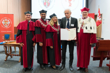 Five people pose for a photo in the auditorium. Four in red and black togas and birettas, one in a suit holding a diploma. In the background are two banners and a screen with the university's logo, on the right a red flag with the coat of arms.