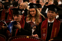 A group of graduates sit in the auditorium. They are wearing black togas and birettas with the emblems of the university. Red sashes read “CLASS OF 2025” and “GRADUATE”. Some are holding programmes or brochures. Other participants in the ceremony can be seen in the background.