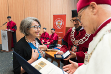 Presentation of medals. A woman receives a medal from a man in academic dress with a rector's chain. Next to him is another man in a toga. In the background people in red togas and birettas and a lectern with the university logo. On the wall a banner with the coat of arms and the inscription Medical University of Warsaw.
