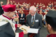 Presentation of medals. A woman receives a medal from a man in academic dress with a rector's chain. Next to him is another man in a toga. In the background people in red togas and birettas and a lectern with the university logo. On the wall a banner with the coat of arms and the inscription Medical University of Warsaw.