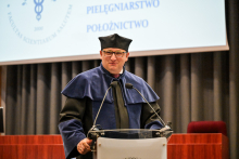 Mężczyzna w okularach, prodziekan Rzońca, w niebiesko-czarnej todze i birecie stoi za mównicą. Przemawia, lekko się uśmiechając. Za nim widać ekran projekcyjny i szare zasłony.
