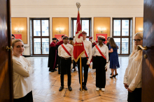 Three students in white shirts and black trousers stand in the middle of a room with a wooden floor. One is holding a red flag with a white eagle. In the background are people in togas and two people at the door. The hall has bright walls, large windows and lamps on the walls.