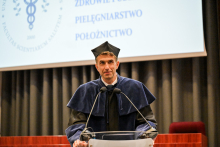 Mężczyzna, prodziekan Raciborski, w niebiesko-czarnej todze i birecie stoi za mównicą. Przemawia, lekko się uśmiechając. Za nim widać ekran projekcyjny i szare zasłony.
