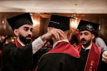 Three graduates in togas and birettas during the ceremony. Two have red sashes with “GRADUATION” written on them. One improves the biretta of the other. In the background, wooden wall panels and lamps.