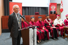 A man in a suit speaks at the lectern with a microphone. In the background, six people in red togas and birettas, two in white togas with rector's chains. Red banners with the university's logo are visible.