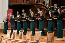 A mixed choir performs in a concert hall. The conductor stands facing the choir with his hands raised. The women are wearing green skirts and black blouses, the men black suits with bow ties. In the background a large instrument and a red banner with the logo “Warsaw Medical University”. Empty wooden chairs in the foreground.
