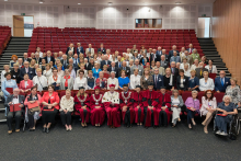 About 100 people pose for a joint photo in the auditorium. In the front row sit people in red academic togas and senior citizens. Behind them stand the other participants against a background of red chairs. Stairs and doors are visible in the background.