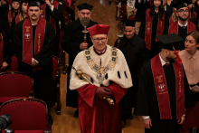 Academic ceremony. In the centre, a person in red dress and white coat with rector's insignia, holds a scepter. To the sides, graduates in black togas and birettas, with maroon “CLASS OF 2025 GRADUATE” sashes. In the background, participants in the ceremony.
