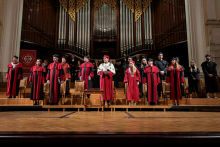 A group of people in red and black togas and birettas stand on stage in the concert hall. In the centre a person in a white toga with a rector's chain. In the background a large silver pipe organ. Wooden floor, decorative architectural elements visible.