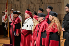 A group of people in academic togas stand on stage. In the foreground is a person in a red toga with white fur and sceptre. The others have red togas with black elements and black birettas. In the background are men in white shirts with flags. The scene is set against a marble wall.