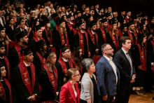 A group of graduates in black togas and birettas stand during the ceremony. They are wearing red stoles with the inscription “CLASS OF 2025”. Seated guests are visible in the background.