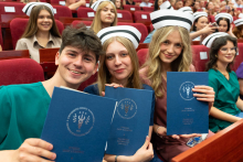Uroczystość czepkowania absolwentów pielęgniarstwa i położnictwa. Młode kobiety i młody mężczyzna w uniformach medycznych i czepkach pielęgniarskich siedzą w audytorium, trzymając niebieskie teczki z napisem „Warszawski Uniwersytet Medyczny”