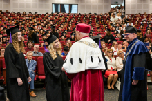 Ceremonia wręczenia dyplomów. Dwie absolwentki w czarnych togach i biretach odbierają dyplomy od mężczyzn w strojach ceremonialnych: czerwonym z białym kołnierzem i niebieskim. W tle sala pełna studentów i gości.