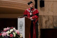 A man in a toga and biretta stands by a rostrum bearing the coat of arms and the inscription ‘Karol Marcinkowski University of Medical Sciences in Poznań’. He is wearing a rector's chain around his neck. In front of the rostrum is a bouquet of pink, white and purple flowers. In the background, a marble wall and a decorative cornice.