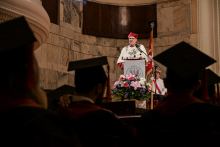 Academic ceremony. In the centre, a speaker in a white robe and red biretta behind a lectern with the university crest. In front of him a bouquet of flowers. In the background a marble wall and a flag. In the foreground, silhouettes of graduates in black togas and birettas, seated backwards.