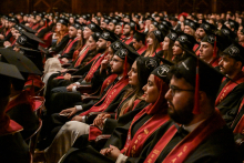 A large group of graduates in black togas and birettas sit in the auditorium. They have red sashes with the words “Class of 2025” written on them. The auditorium is full. In the background, the audience is visible in the balconies.