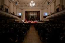 A large concert hall with a high ceiling and chandelier. On stage, people in red togas sit on chairs. A large pipe organ in the background. On the sides of the stage, screens showing the person speaking at the lectern. The audience sits in darkness.