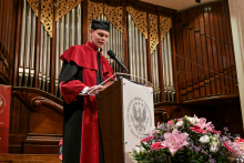 A man in a toga and biretta stands behind a lectern with the logo of the Medical University of Warsaw. In front of him is a microphone and a bouquet of flowers in shades of pink, white and red. In the background, marble walls and wooden panels.