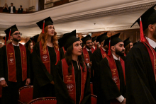A group of students in black togas and birettas stand in rows during the graduation ceremony. The red sashes read ‘CLASS OF 2025’. In the background is an elegant hall with a balcony where guests are seated.