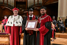 Three men in academic togas stand on stage. The middle one holds a diploma. On the left a person in a red toga with white fur and chain. On the right, a person in a red and black toga with a chain. In the background, participants in the ceremony in togas. Stage in an elegant interior with wood and pipe organ.