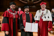 Three men in togas and birettas stand against a wooden wall. The middle one in a black toga with a maroon “Class of 2025 Graduate” sash holds a diploma. To the sides, people in red and red and white togas with rector's chains. In the background, other participants in the ceremony.