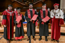 Five people in togas and birettas stand on stage. Three people in the centre hold red diplomas. To the sides, people in red togas with decorative chains. In the background, wooden decoration and seated ceremony participants.