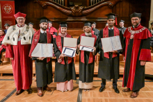 A group of graduates in black togas and birettas pose with their diplomas. To the sides stand people in red togas with rector's insignia. In the background, the wooden decoration of the hall and the organ.