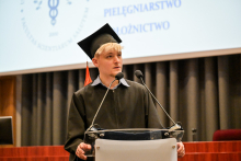 Młody mężczyzna z jasnymi włosami w czarnej todze i birecie przemawia na podium. Biret ma chwost w kolorze pomarańczowym. W tle widać szare zasłony i ekran projekcyjny.