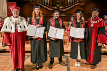 Six people in togas and birettas stand on the wooden floor. Four people in the middle have black togas with red sashes and are holding diplomas. To the sides, people in red togas with white elements and chains. In the background, a wooden organ and a red plaque with the university's coat of arms.